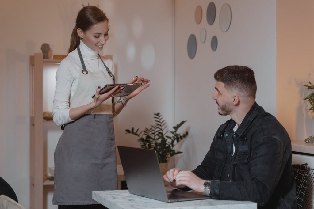A smiling waitress takes an order from a customer on a tablet in a cozy café setting.